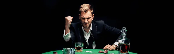 Panoramic shot of angry man with clenched hand near alcohol and playing cards isolated on black — Stock Photo