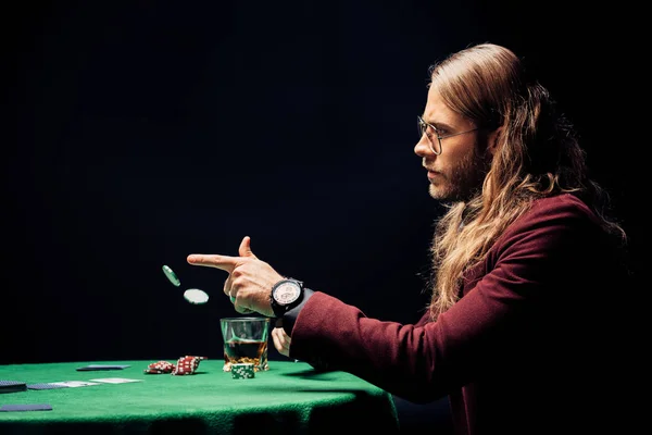 Vista lateral do homem em óculos de olho perto de vidro com uísque jogando em fichas de poker ar isolado em preto — Fotografia de Stock