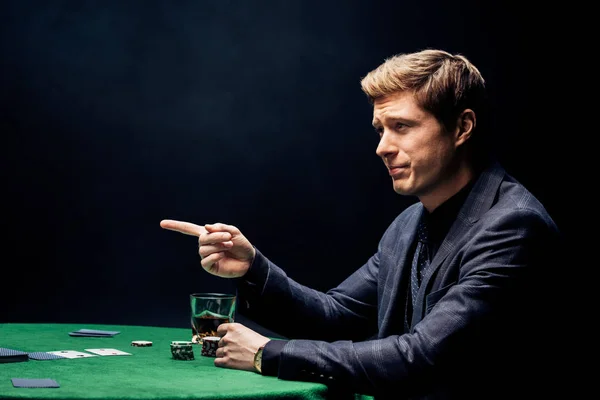 Handsome man pointing with finger near poker table on black — Stock Photo