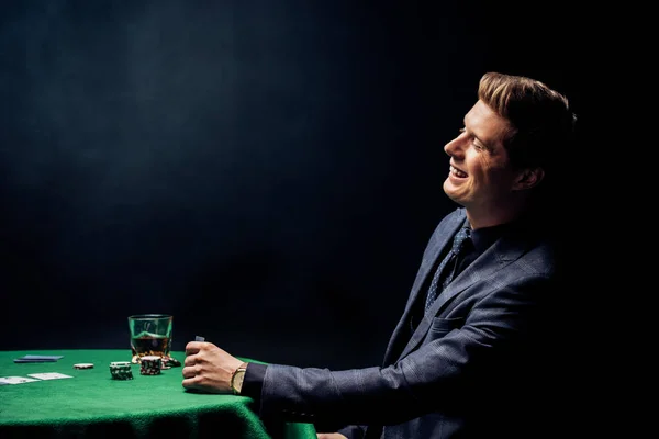 Cheerful man smiling near poker table with glass of whiskey on black — Stock Photo