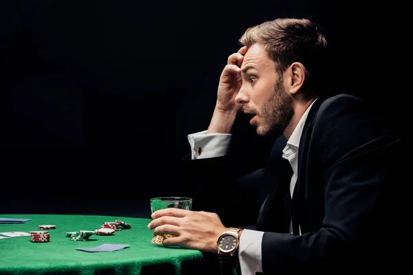 QUIIV, UCRÂNIA - 20 DE AGOSTO DE 2019: vista lateral de um homem surpreso jogando poker perto de um copo de uísque isolado em preto — Fotografia de Stock