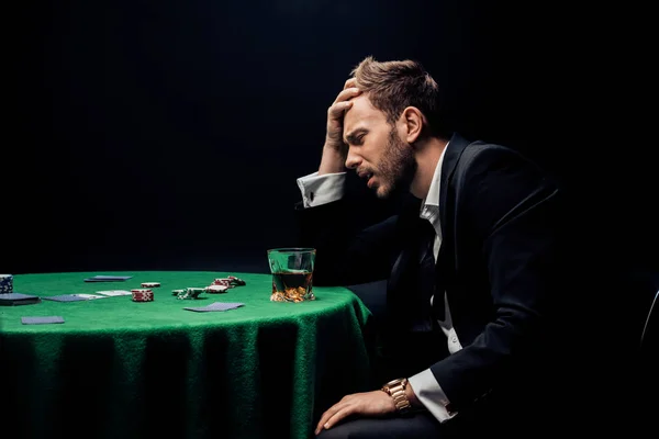 KYIV, UKRAINE - AUGUST 20, 2019: side view of sad man playing poker near glass of whiskey isolated on black — Stock Photo