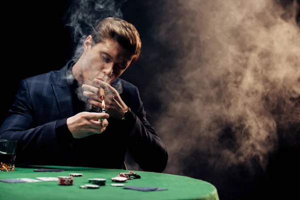 Selective focus of handsome man holding lighter while smoking on black with smoke — Stock Photo