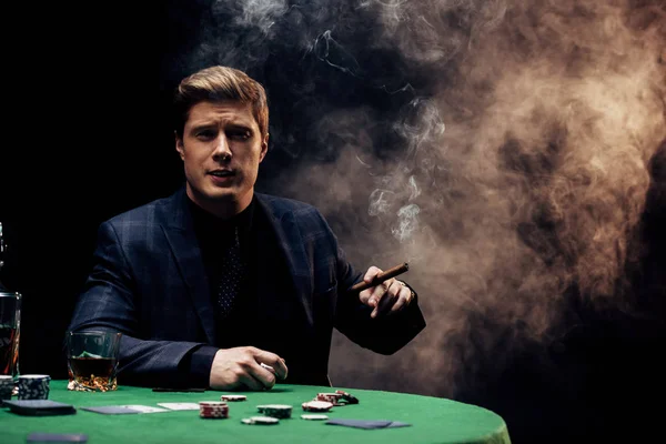 Handsome man holding cigar while smoking on black with smoke — Stock Photo