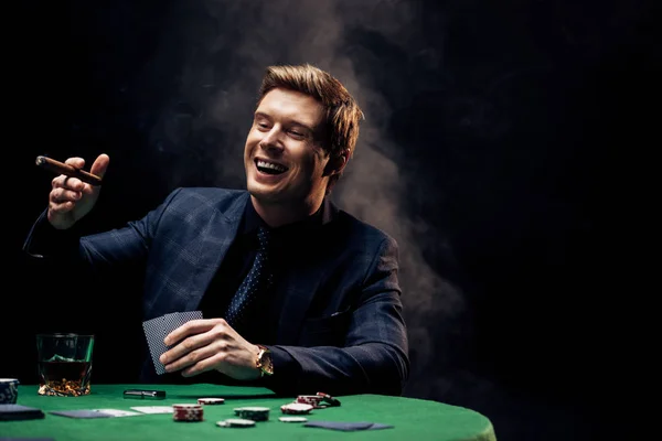 KYIV, UCRANIA - 20 de agosto de 2019: hombre feliz sosteniendo cigarros y jugando a las cartas en negro con humo — Stock Photo