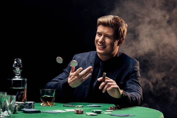 Cheerful man throwing in air poker chips and cigar on black with smoke — Stock Photo
