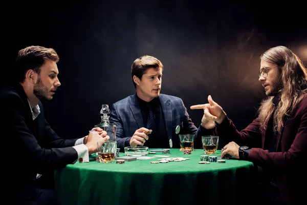Hombre señalando con el dedo cerca de amigos mientras juega al póquer en negro con humo - foto de stock