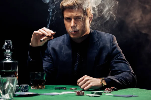 Handsome man smoking cigar near poker table on black with smoke — Stock Photo
