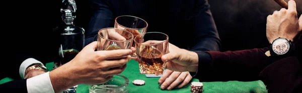 Panoramic shot of men clinking glasses with whiskey on black — Stock Photo