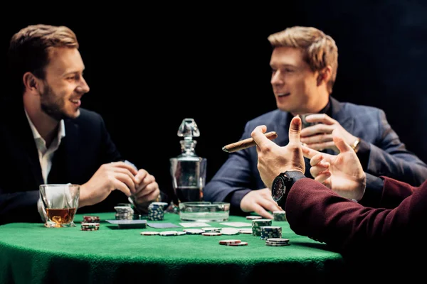 Cropped view of man holding cigar near friends in poker club on black — Stock Photo