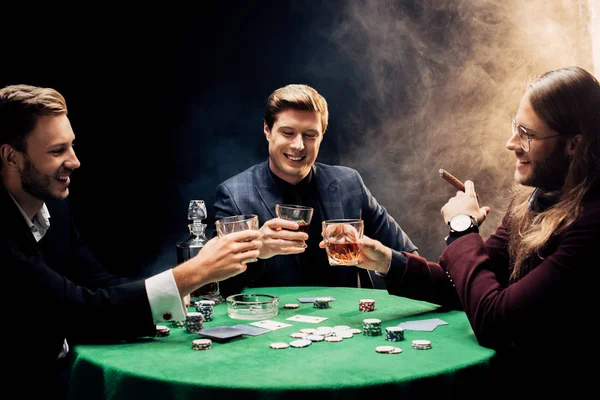 KYIV, UKRAINE - AUGUST 20, 2019: happy men holding glasses with alcohol near poker table on black with smoke — Stock Photo