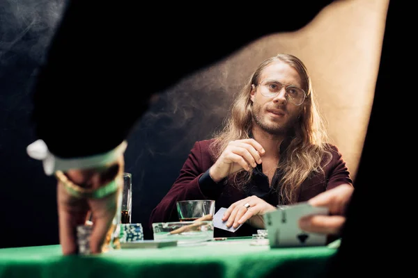 KYIV, UKRAINE - AUGUST 20, 2019: selective focus of handsome bearded man in eye glasses playing poker with friend on black with smoke — Stock Photo