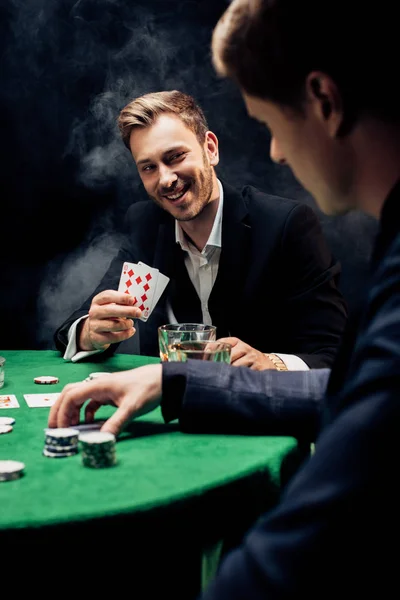 KYIV, UKRAINE - 20 AOÛT 2019 : mise au point sélective de l'homme heureux jouant au poker ami proche et jetons de poker sur noir avec de la fumée — Photo de stock