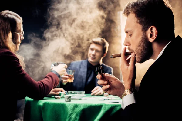 Selective focus of handsome man smoking while playing poker on black with smoke — Stock Photo