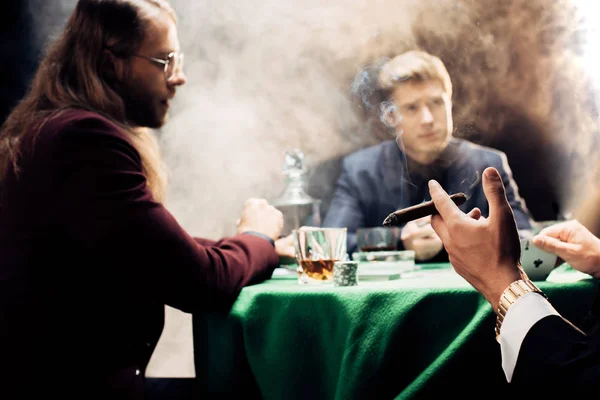 Vue recadrée de l'homme tenant cigare tout en jouant au poker sur noir avec de la fumée — Stock Photo