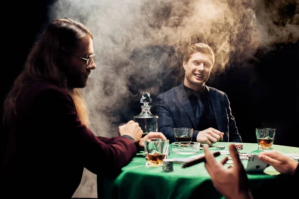 KYIV, UCRANIA - 20 de agosto de 2019: vista recortada del hombre sosteniendo cartas cerca de jugadores felices en negro con humo - foto de stock