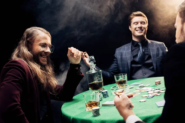 Vista recortada del hombre cerca de amigos felices cerca de la mesa de póquer en negro con humo - foto de stock