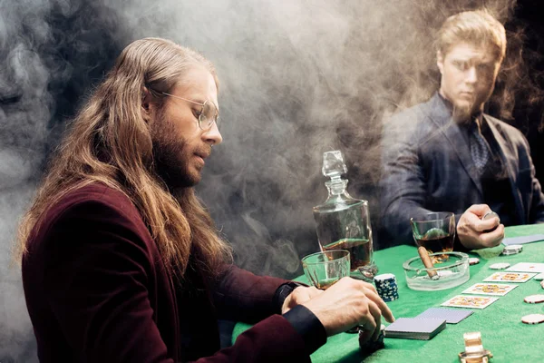 KYIV, UKRAINE - AUGUST 20, 2019: handsome men playing poker near poker table with poker chips and playing cards on black with smoke — Stock Photo