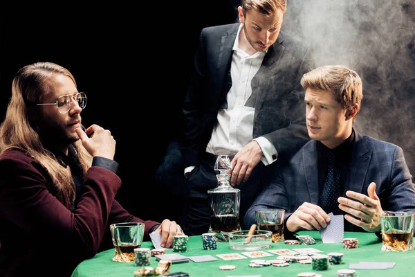 KYIV, UKRAINE - AUGUST 20, 2019: handsome men gesturing while playing poker on black with smoke — Stock Photo