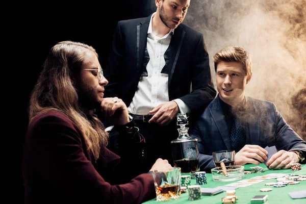 KYIV, UKRAINE - AUGUST 20, 2019: handsome men playing poker on black with smoke — Stock Photo