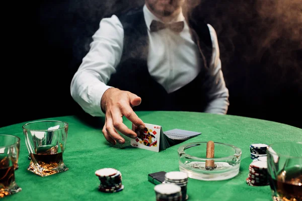 КИЕВ, УКРАИНА - 20 августа 2019 года: обрезанный вид крупье касаясь игральных карт возле покерного стола на черном с дымом — стоковое фото