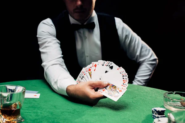 Київ, Україна - 20 серпня 2019: Переглянутий погляд на кур'єр у формальному носі, що тримає гральні карти біля покерного столика ізольованого на чорному — Stock Photo