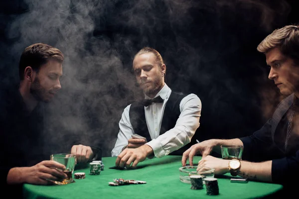 KYIV, UKRAINE - AUGUST 20, 2019: croupier holding playing cards near men on black with smoke — Stock Photo