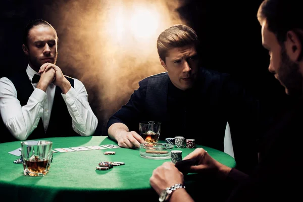 KYIV, UKRAINE - AUGUST 20, 2019: handsome men playing poker near croupier on black with smoke — Stock Photo