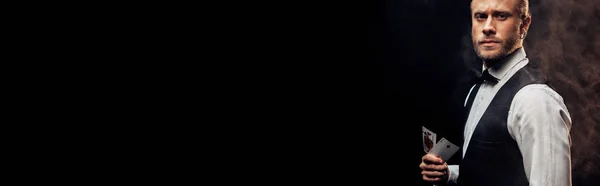 KYIV, UCRANIA - 20 de agosto de 2019: tiro panorámico croupier barbudo guapo sosteniendo cartas en negro con humo - foto de stock