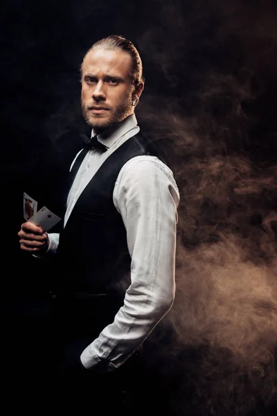 KYIV, UKRAINE - AUGUST 20, 2019: handsome croupier holding playing cards on black with smoke — Stock Photo