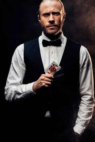 KYIV, UKRAINE - AUGUST 20, 2019: handsome bearded croupier holding playing card on black with smoke — Stock Photo