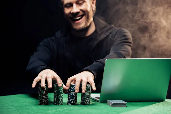 KYIV, UCRANIA - 20 de agosto de 2019: hombre feliz tocando fichas de póquer cerca del ordenador portátil en negro con humo - foto de stock