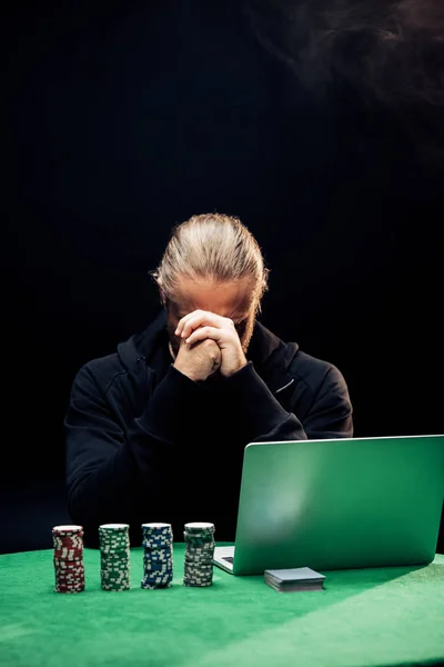 KYIV, UKRAINE - AUGUST 20, 2019: man covering face while using laptop near poker chips on black with smoke — Stock Photo