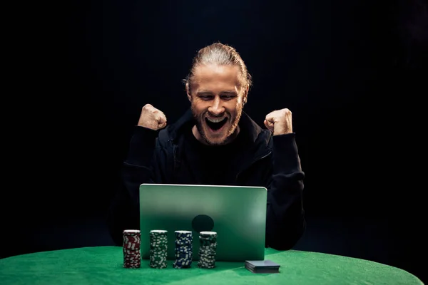 KYIV, UKRAINE - 20 de agosto de 2019: homem feliz gesticulando ao usar laptop perto de fichas de poker isoladas em preto — Fotografia de Stock