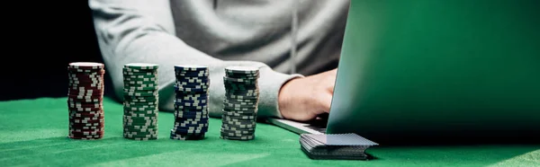 KYIV, UCRANIA - 20 de agosto de 2019: plano panorámico del hombre usando el ordenador portátil cerca de fichas de póquer aisladas en negro - foto de stock