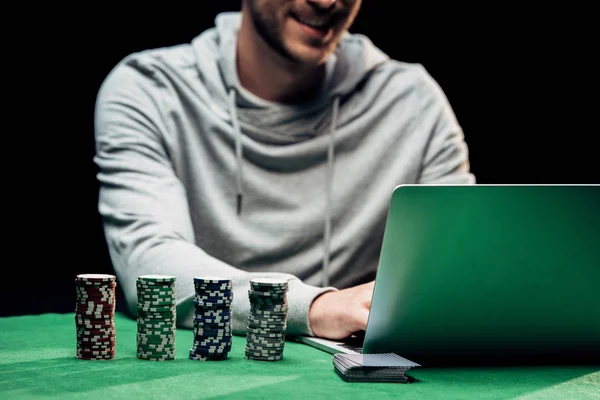 KYIV, UKRAINE - 20 de agosto de 2019: visão recortada do homem feliz usando laptop perto de fichas de poker isoladas em preto — Fotografia de Stock