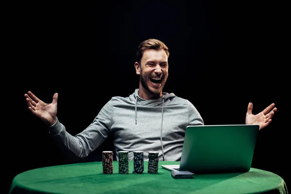 Kyiv, Ukraine - 20. August 2019: selektiver Fokus des glücklichen Mannes gestikuliert in der Nähe von Pokerchips in der Nähe von Laptop isoliert auf schwarz — Stockfoto