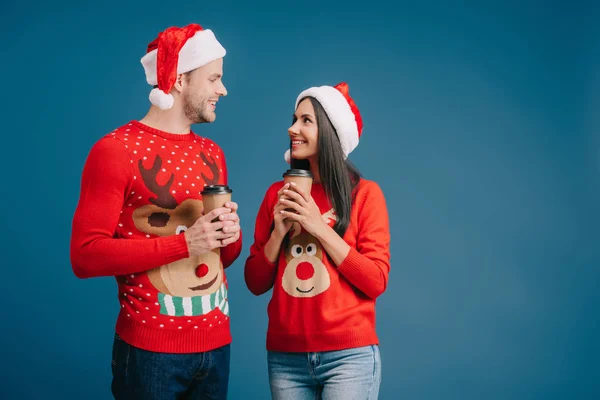 Beau couple en chapeaux de Père Noël et pulls de Noël tenant café pour aller isolé sur bleu — Photo de stock
