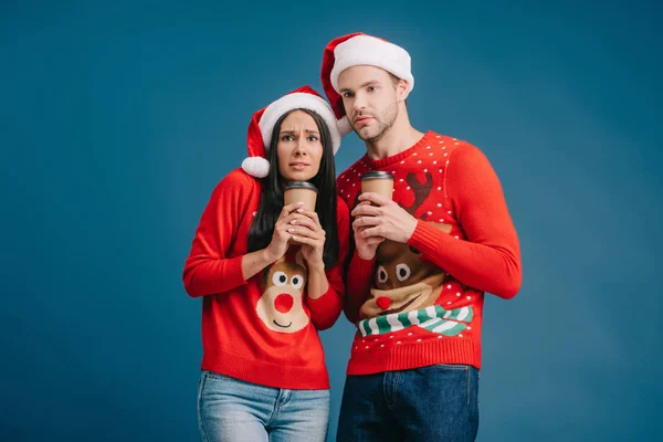Cold couple in santa hats and christmas sweaters holding coffee to go isolated on blue — Stock Photo