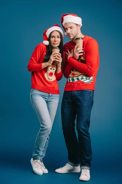 Hermosa pareja fría en sombreros de santa y suéteres de Navidad celebración de café para ir aislado en azul - foto de stock
