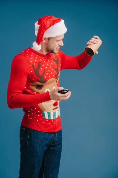 Hombre en sombrero de santa y suéter de Navidad sosteniendo taza vacía de café, aislado en azul - foto de stock