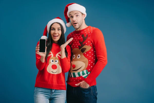 Здивована пара в капелюхах Санти і різдвяних светрах, що показують смартфон з порожнім екраном ізольовано на синьому — стокове фото