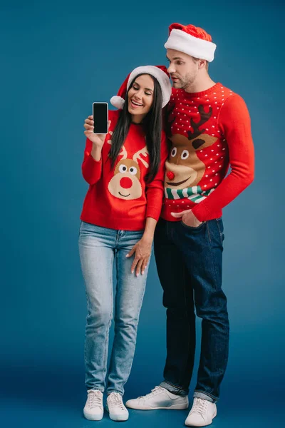Fröhliches Paar in Weihnachtsmützen und Weihnachtspullovern zeigt Smartphone mit leerem Bildschirm isoliert auf blauem Grund — Stockfoto