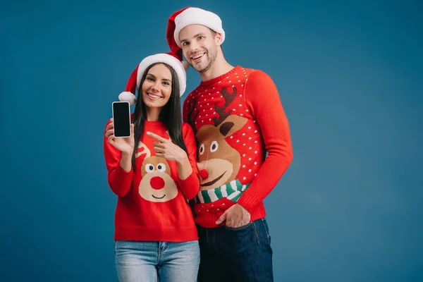 Пара в шляпах Санта-Клауса и рождественских свитерах, указывающих на смартфон с пустым экраном, изолированным на синем — стоковое фото