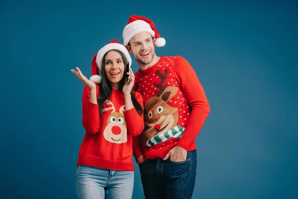 Glückliche Frau mit Weihnachtsmannmütze, die auf dem Smartphone spricht, während Mann zuhört, isoliert auf blauem Grund — Stockfoto