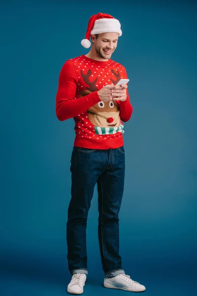 Красивий чоловік у капелюсі Санта та різдвяному светрі за допомогою смартфона, ізольований на синьому — стокове фото