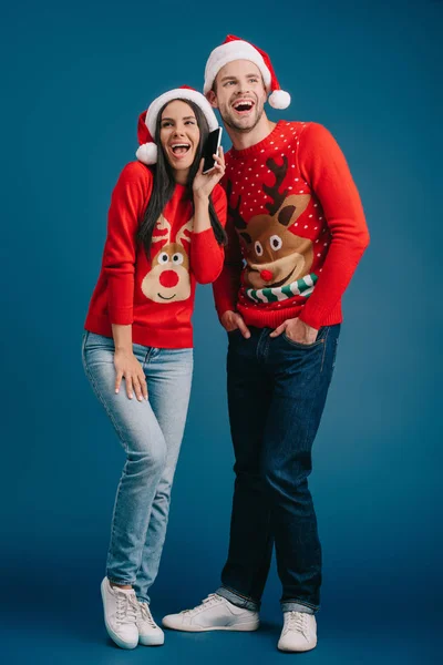 Pareja riendo en sombreros de santa y suéteres de Navidad hablando teléfono inteligente aislado en azul - foto de stock