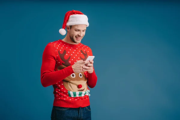 Усміхнений чоловік у капелюсі Санти та різдвяному светрі за допомогою смартфона, ізольований на синьому — стокове фото
