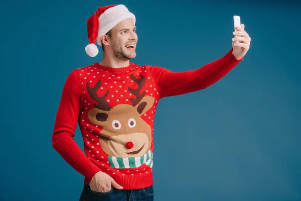 Красивий щасливий чоловік в капелюсі Санта і різдвяний светр, приймаючи селфі на смартфон, ізольований на синьому — стокове фото
