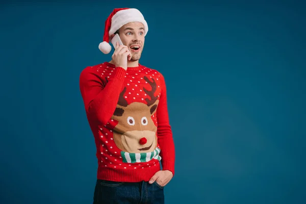 Uomo eccitato in cappello di Babbo Natale e maglione di Natale parlando su smartphone, isolato sul blu — Foto stock